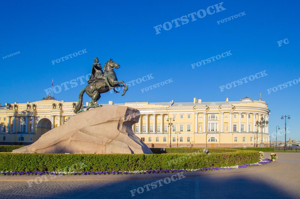 На фотографии изображен Медный всадник — знаменитый памятник Петру I на фоне здания Сената и Синода в Санкт-Петербурге. Памятник расположен на Сенатской площади, это один из символов города. Небо ясное, что указывает на ясный день, возможно, утро, учитывая направление тени. Фотография может использоваться для туристических проспектов, исторических книг, образовательных материалов и как реклама для посещения города.